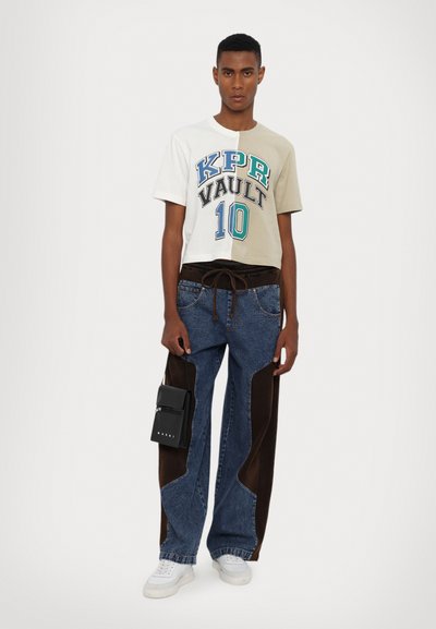 T-shirt accorciato in beige chiaro e bianco con testo grafico; pantaloni in denim blu con accenti in suede marrone; tiene una piccola borsa nera.