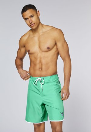 Fit man adjusting light green swim shorts with white drawstring and trim on plain light gray background.