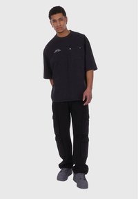 Sort oversized T-shirt med en brystlomme og hvid logo detalje, sammen med sorte cargo bukser med sidelommer og grå sko.