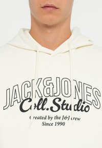 Hvid hættejakke med et stort sort tekstdesign, der står "JACK & JONES Coll. Studio". Den har en snore-træk hætte og en blød stofstruktur.