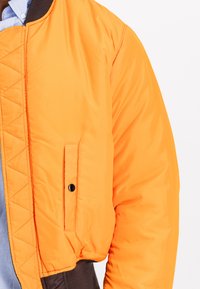 Oranžová prešívaná bomber bunda s čiernym rebrovaným golierom a pasom. Obsahuje vrecká so čiernymi zapínaním na cvoky a hladkým, lesklým materiálom.