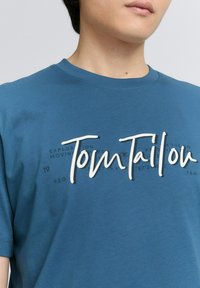 Mėlynas medvilninis marškinėliai su dideliu baltu "Tom Tailor" logotipu ir mažesniu tekstu priekyje. Trumpomis rankovėmis ir apvaliu kaklu.