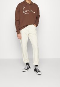 Felpa marrone con colletto a zip e logo bianco, abbinata a pantaloni crema tapered e sneakers alte nere.
