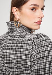 Top din material în carouri alb-negru, cu guler ondulat; accesorii cu cercei mari din aur, cu detalii intricate.