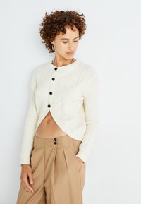 Cardigan crème en maille torsadée avec un design court, doté de boutons noirs sur le devant et de poignets côtelés, associé à un pantalon beige plissé.