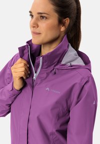 Giacca impermeabile viola con colletto alto, zip frontale e dettagli di ventilazione. Logo posizionato sul lato sinistro del petto e sulla spalla.