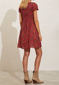 Kortärmad klänning i magenta med intrikat paisleymönster, med en rynkad överdel och en utsvängd kjol. Bärs med beigea ankelstövlar.