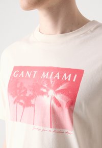 Krémové bavlněné tričko s růžovým grafickým motivem palem a textem "GANT MIAMI" a pod ním v písmu "Pozdravy ze Slunného státu".