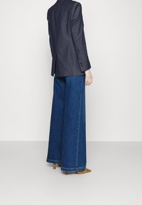 Tmavý blazer s přiléhavým střihem, v kombinaci se širokými modrými džíny. Výrazné prošívání na manžetách a vzorovaná obuv.