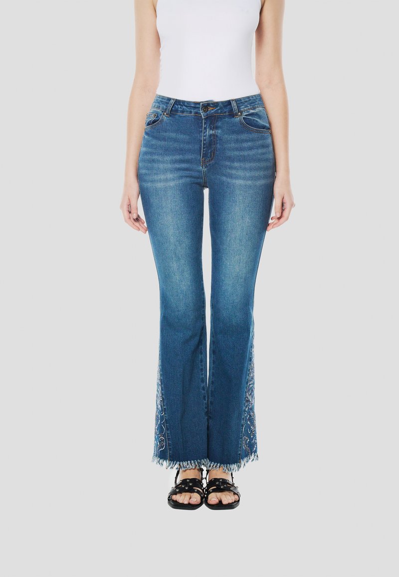 Közép kék mosású, bővülő szárú farmernadrág rojtos szegéllyel és oldalsó virágos hímzéssel. Elöl zsebekkel és gombos záródással rendelkezik.