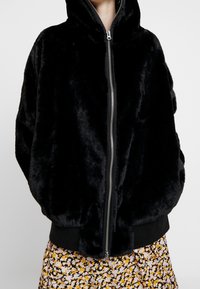 Czarna kurtka z sztucznego futra z kapturem, z pełnym zamkiem błyskawicznym i ściągaczami przy mankietach. Teksturowany, oversize'owy fason; noszona na spódnicę w kwiatowy wzór.