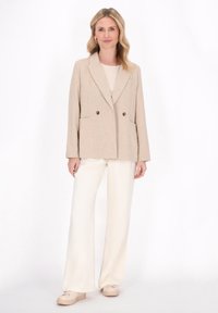 Blazer beige doppiopetto in tessuto testurizzato con tasche frontali, abbinato a pantaloni a gamba larga crema e sneakers di tonalità chiara.