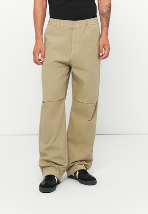 Man wearing beige wide-leg pants and black sneakers with white stripes, standing against a plain white background.