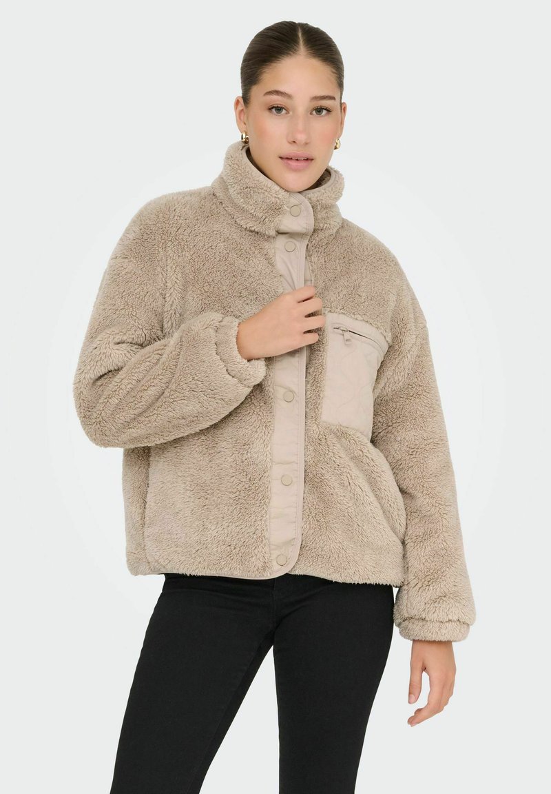 Beżowa kurtka z imitacji futra z wysokim kołnierzem, wyposażona w kieszeń na zamek błyskawiczny oraz zapięcie na zatrzask. Miękka w dotyku i o luźnym kroju.