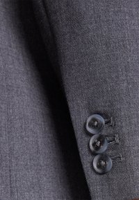 Detalle de chaqueta de traje gris que muestra tela texturizada, tres botones transparentes en el puño y costuras entalladas.