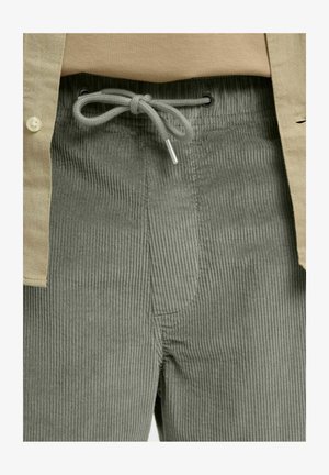 Nærbilde av olivengrønne kordfløyelbukser med knytt midjesnor og del av en beige skjorte med knapper synlig.