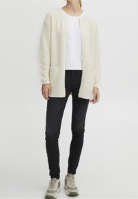 Kardigan o kremowej fakturze z otwartym przodem, z dwiema kieszeniami. W zestawie z białym t-shirtem, czarnymi dopasowanymi dżinsami i szarymi sneakersami.