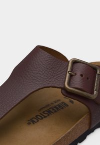 Καφέ δερμάτινο σανδάλι Birkenstock με ρυθμιζόμενο ορείχαλκο αγκράφα και ανάγλυφη φελλό σόλα, εμφανές σε λευκό φόντο.
