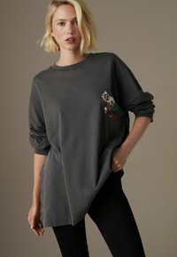 Übergroßes graues Sweatshirt aus Baumwolle, mit einem bunten gestickten Design auf der linken Brustseite und aufgeschlagenen Bündchen.