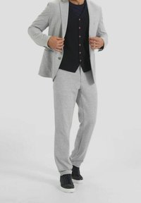 Uomo che indossa un completo grigio chiaro con pantaloni abbinati, un gilet nero con bottoni, una camicia blu navy e sneakers nere con suola bianca.