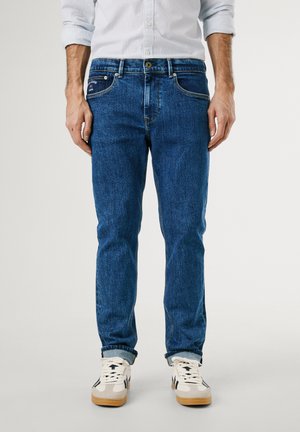 Jean en denim bleu à coupe droite, avec des coutures jaunes, cinq poches et une fermeture à boutons. Porté avec des baskets blanches.