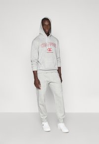 Γκρι hoodie με το λογότυπο "CHAMPION" σε κόκκινο, κουκούλα με κορδόνια, μακριά μανίκια. Γκρι φούτερ παντελόνια με ελαστικά τελειώματα. Λευκά αθλητικά παπούτσια.