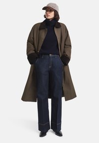 Palton lung verde măsliniu cu guler maro închis, pulover de culoare navy cu guler înalt, pantaloni largi din denim închis cu cusături în contrast și cizme negre cu toc.