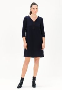 Marineblaues Kleid mit Dreiviertelärmeln, V-Ausschnitt, Reißverschlussdetail vorne und lockerer Passform. Kombiniert mit schwarzen Knöchelstiefeln.