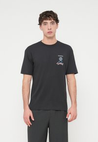 Μαύρο βαμβακερό t-shirt με κοντά μανίκια, με λογότυπο Toyota και γραφιστικό σχέδιο στο στήθος, σε συνδυασμό με σκούρο γκρι παντελόνι.