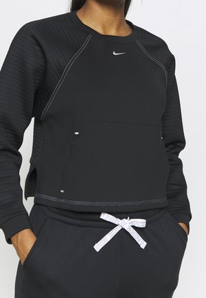 Vrouw draagt een zwarte Nike sweater en bijpassende broek met wit Nike-logo op de borst en wit trekkoord gestrikt ter hoogte van de taille.