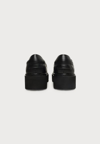 Paire de chaussures à plateforme en cuir noir vues de dos, avec semelles épaisses et coutures visibles sur un fond clair uni.