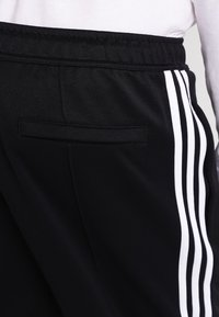 Černé sportovní kalhoty s elastickým pasem, zadním klínovým kapsou a třemi bílými svislými pruhy na boku.