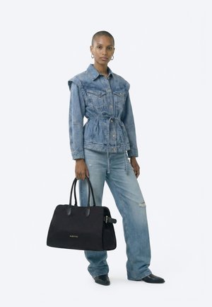 Žena oblečená v modré džínové bundě s páskem a džínách, držící velkou černou kabelku Valentino, stojící na bílém pozadí.