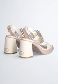 Crème kleurige hoge hakken sandalen met een decoratief uitsnijdingsontwerp, dikke blokhakken en accenten in goudkleurige hardware.