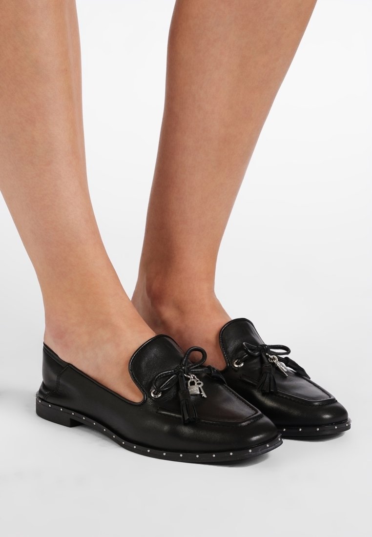 Mocassins en cuir noir avec des pampilles et de petits charmes en forme de serrure et de clé en argent, portés pieds nus sur un fond blanc uni.