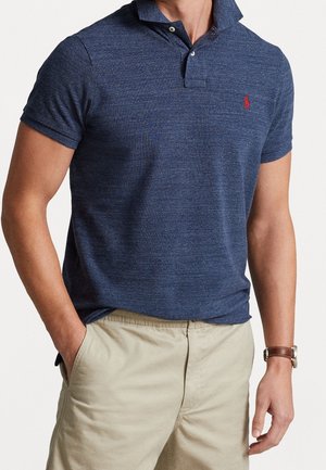 Mężczyzna ubrany w granatową koszulkę polo z krótkim rękawem i czerwonym logo oraz beżowe spodnie, jedna ręka w kieszeni, na białym tle.