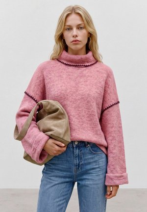 Blond vrouw in een roze coltrui met een donkere rand en blauwe spijkerbroek houdt een beige suède tas vast tegen een effen achtergrond.
