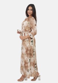 Lange, bloemenjurk in beige met zachte bruine en groene accenten. Heeft een strik taille, vloeiende mouwen en een geplooide rok. Transparante haksandalen.
