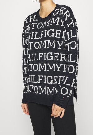 Czarny dziergany sweter z powtarzającym się białym tekstem "TOMMY HILFIGER", noszony przez osobę w czarnych spodniach na jednolitym tle.