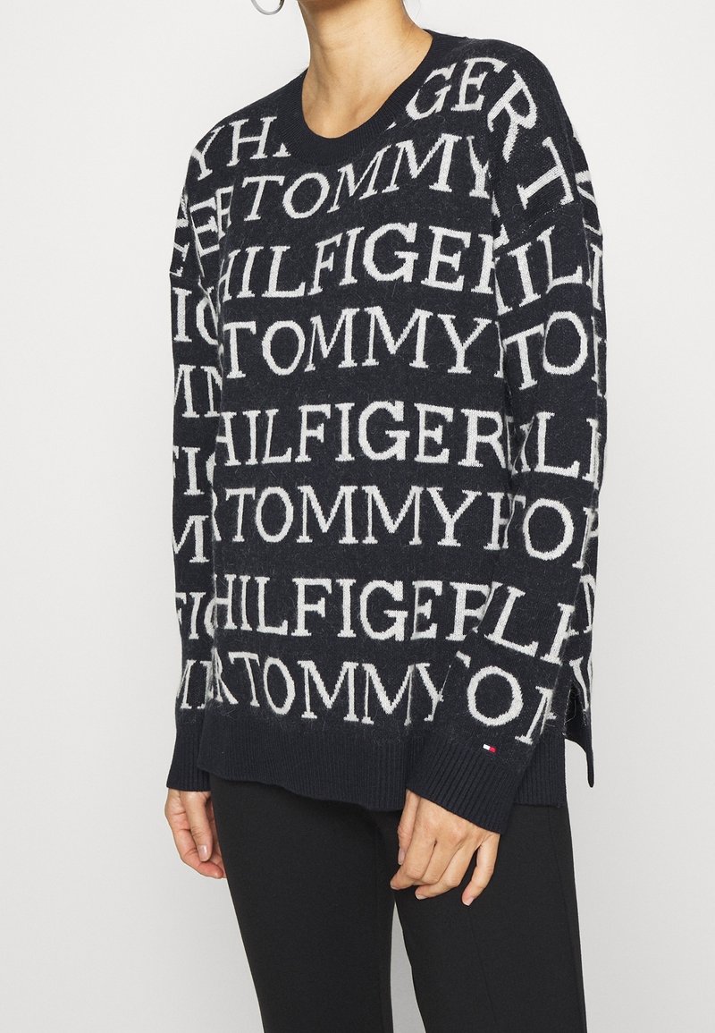 Pull en laine noir avec texte blanc répété "TOMMY HILFIGER", porté par une personne en pantalon noir sur fond uni.