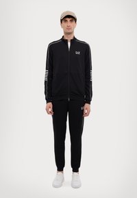 Completo sportivo nero con strisce bianche sulle maniche e sul lato dei pantaloni, composto da una giacca con zip e polsini elasticizzati. Indossato con sneaker bianche.