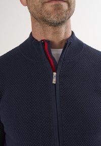 Marineblauer Strick-Zip-Pullover mit strukturiertem Wabenmuster, ausgestattet mit einem hohen Kragen und kontrastierendem roten Reißverschlussband. Minimalistische Designdetails.