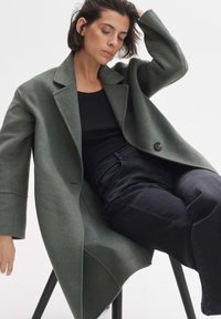 Übergroßer, grüner Wollmischungsmantel mit Reverskragen und einem einzelnen Knopf. Getragen über einem schwarzen Oberteil, kombiniert mit schwarzen engen Jeans.