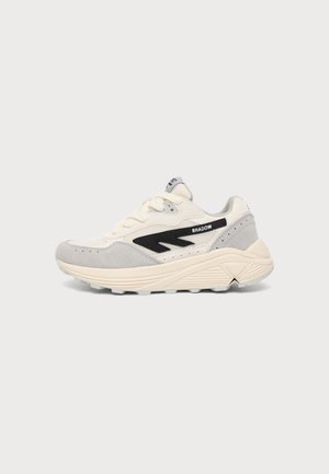 Crème- en lichtgrijze sneaker met dikke zool, zwart zigzagpatroon en "SHADOW" tekst aan de zijkant, weergegeven op een witte achtergrond.