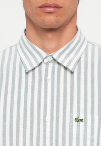 Camisa de algodón a rayas en blanco y verde, con collar abotonado y un pequeño logo bordado en el lado izquierdo del pecho.