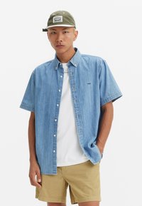 Light blue denim short-sleeve shirt with a button-down collar, worn over a white t-shirt, paired with beige shorts, posing against a white background.