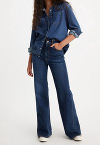 Chemise en denim bleu à boutons avec deux poches poitrines, manches retroussées, associée à un jean évasé bleu foncé et des baskets blanches.