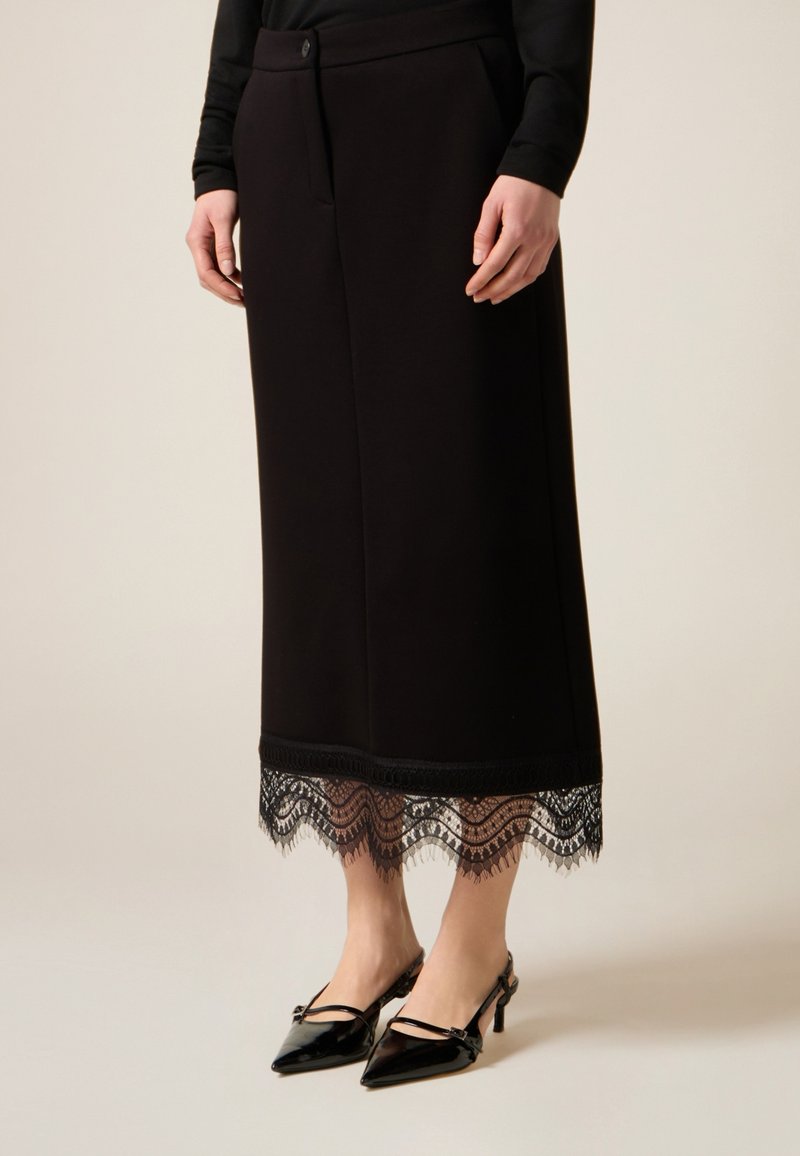 Falda midi negra con un dobladillo de encaje y un patrón detallado. Cuenta con cierre frontal de botones y bolsillos laterales, combinada con tacones negros.