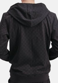 Hombre con una sudadera negra con un sutil patrón geométrico, visto desde atrás contra un fondo claro y liso.