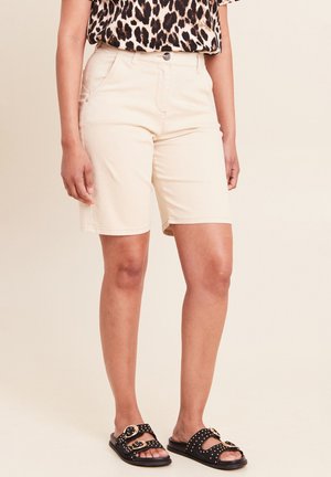 Frau trägt beige knielange Shorts, Leopardenmuster-Top und schwarze Sandalen mit goldenen Schnallen und Nieten, steht vor neutralem Hintergrund.